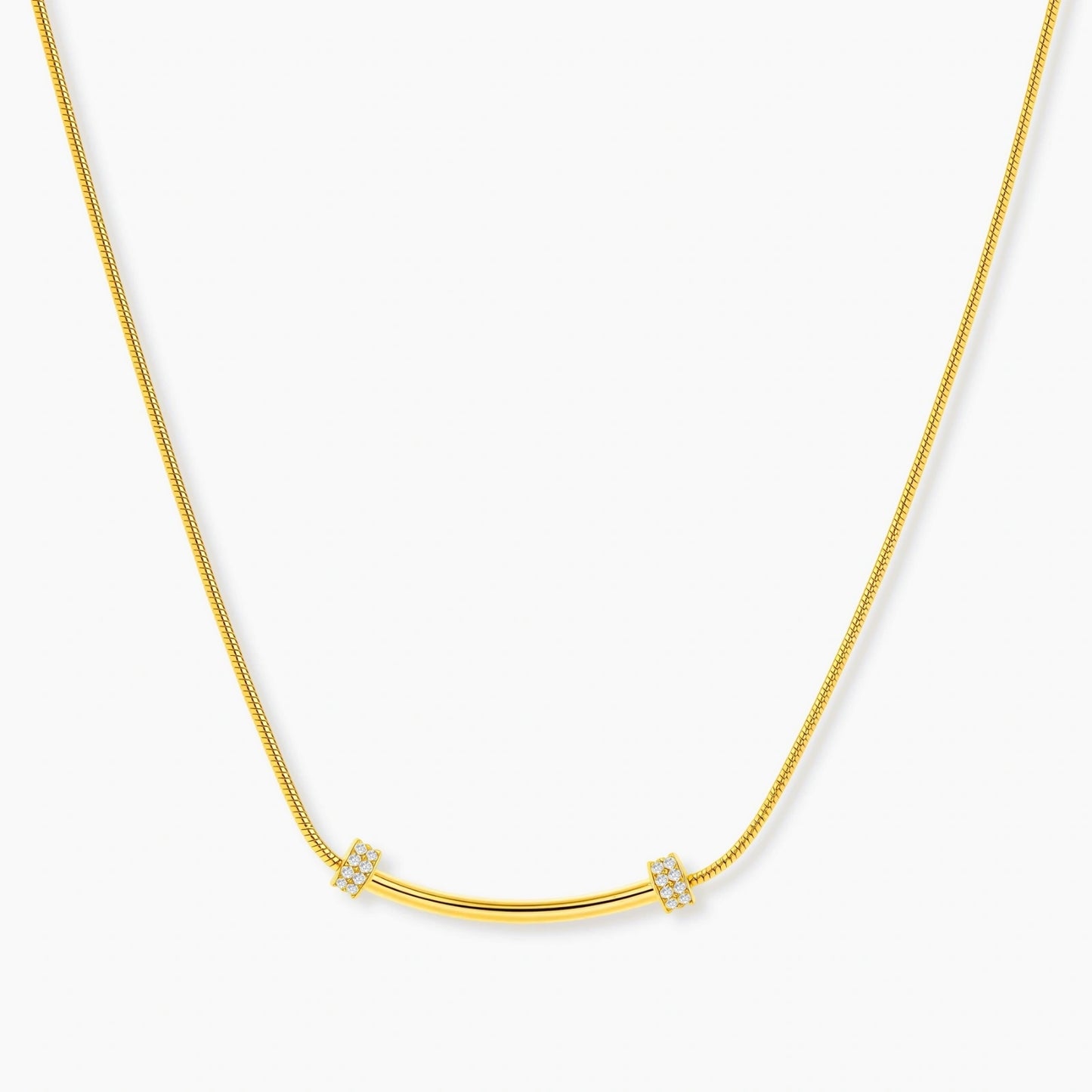 Delicate gold-plated necklace with curved bar pendant and crystal accents on snake chain against cream background.